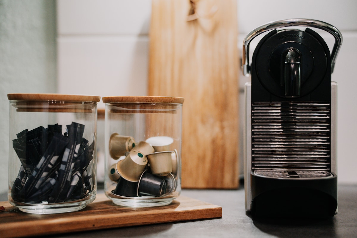 An elegant Nespresso coffee machine is positioned alongside two glass jars. One jar contains various coffee capsules, while the other holds sugar packets. A wooden cutting board provides a natural contrast to the modern kitchen design.