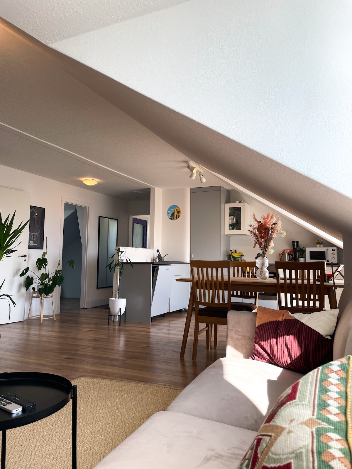 A bright living area is presented featuring a comfortable sofa and a dining table with wooden chairs. A kitchen is visible in the background, equipped with modern appliances. Natural light fills the space, enhanced by warm, neutral tones and decorative plants.
