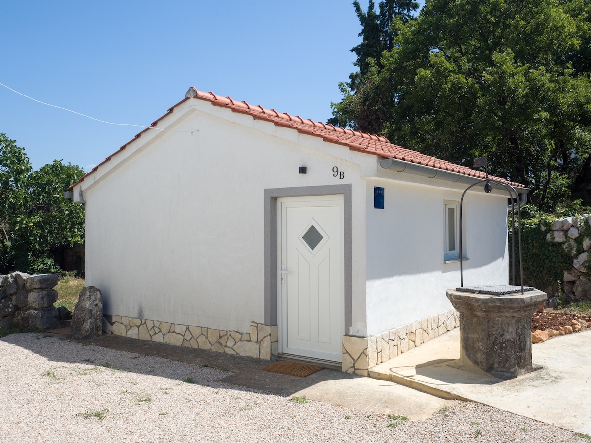 A simple white cottage features a terracotta tile roof and a diamond-shaped window above the door. A stone well stands beside the entrance, set against a backdrop of trees and greenery, creating a peaceful outdoor space.
