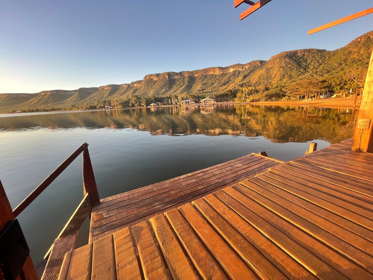 A wooden pier extends over calm waters, reflecting the surrounding mountains and clear sky. The image captures a tranquil view, inviting relaxation. The wooden surface of the pier creates a natural aesthetic, blending harmoniously with the serene setting.