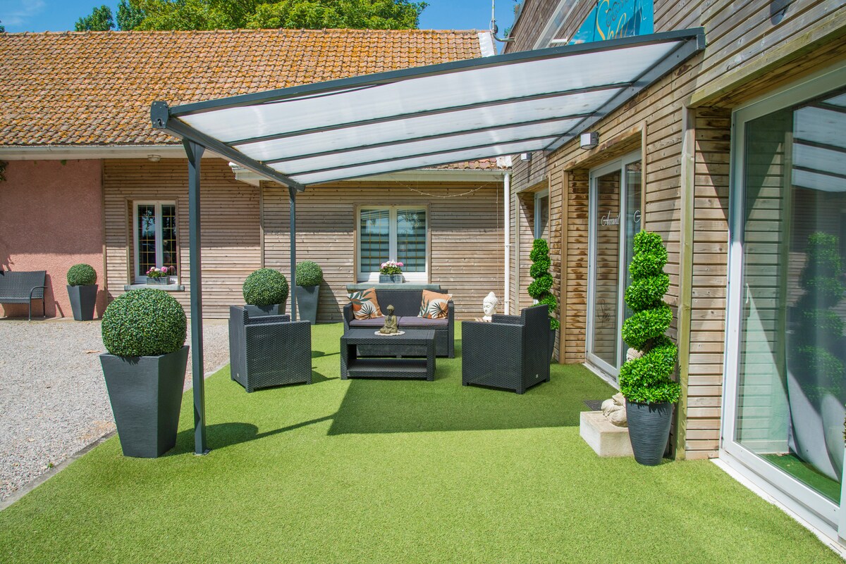 An outdoor seating area is featured, adorned with black wicker furniture and complemented by round topiary plants in sleek planters. A covered terrace provides shade, while artificial grass enhances the inviting space. Large windows of the nearby building reflect the surrounding greenery.