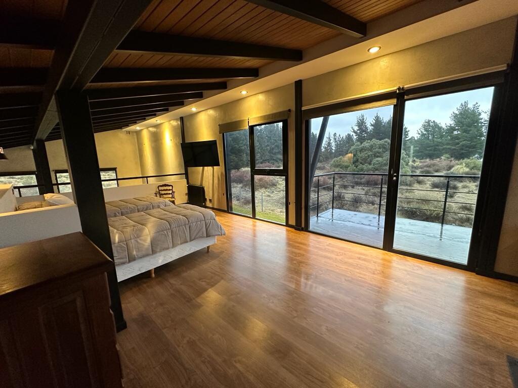 A spacious loft-style bedroom features large glass doors that provide an expansive view of the surrounding nature. The area is illuminated by soft lighting and includes a king-sized bed with neutral bedding. A wall-mounted television is visible, enhancing the functional layout.