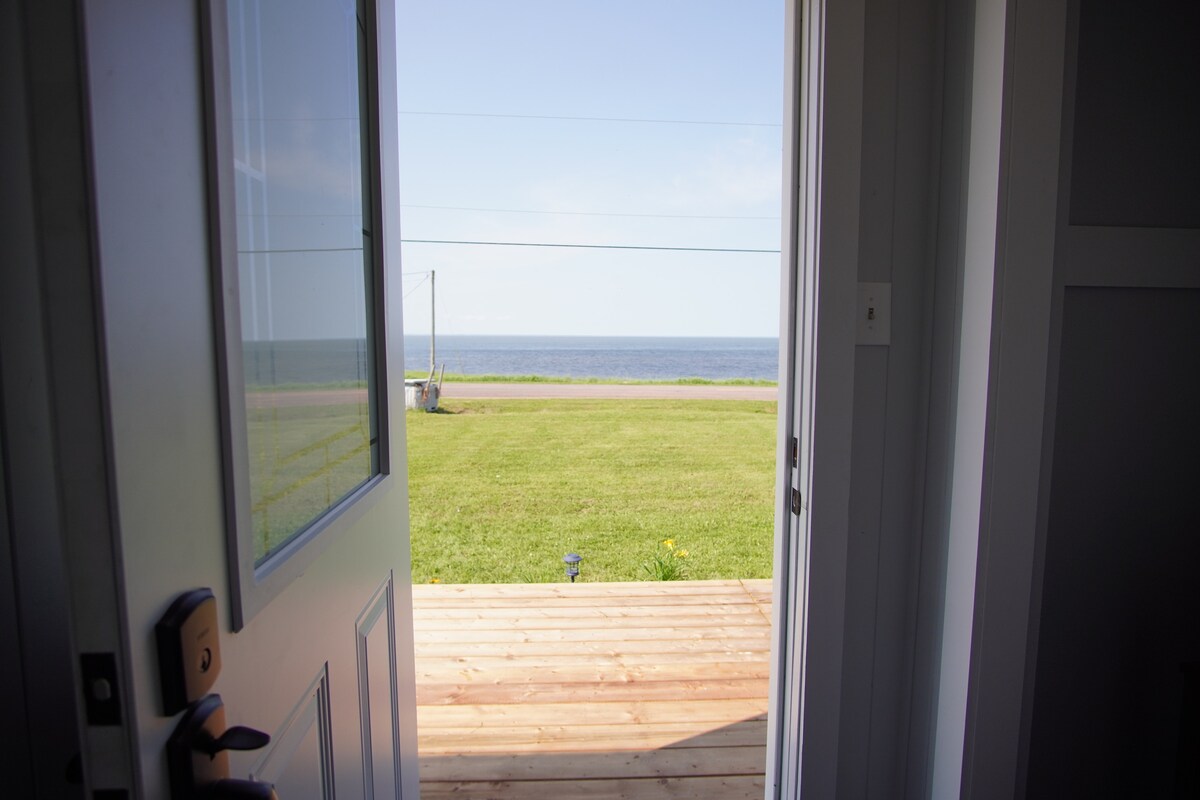 A door stands open, revealing a wooden deck that leads to a grassy area overlooking the ocean. The horizon stretches in the distance, where the sky meets the sea. Gentle sunlight illuminates the scene, highlighting the expansive view.