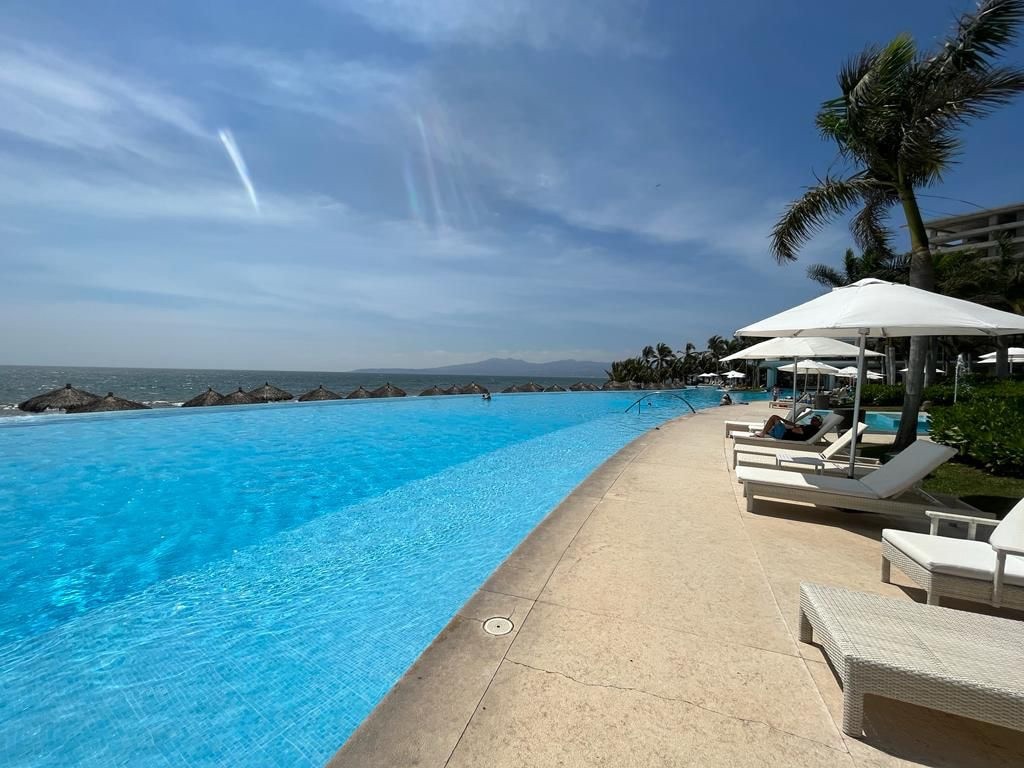 An infinity pool stretches alongside the coastline, blending seamlessly with the ocean. Sun loungers are positioned under white umbrellas, providing shade. Lush greenery borders the area, and thatched umbrellas can be seen in the distance, enhancing the serene water view.