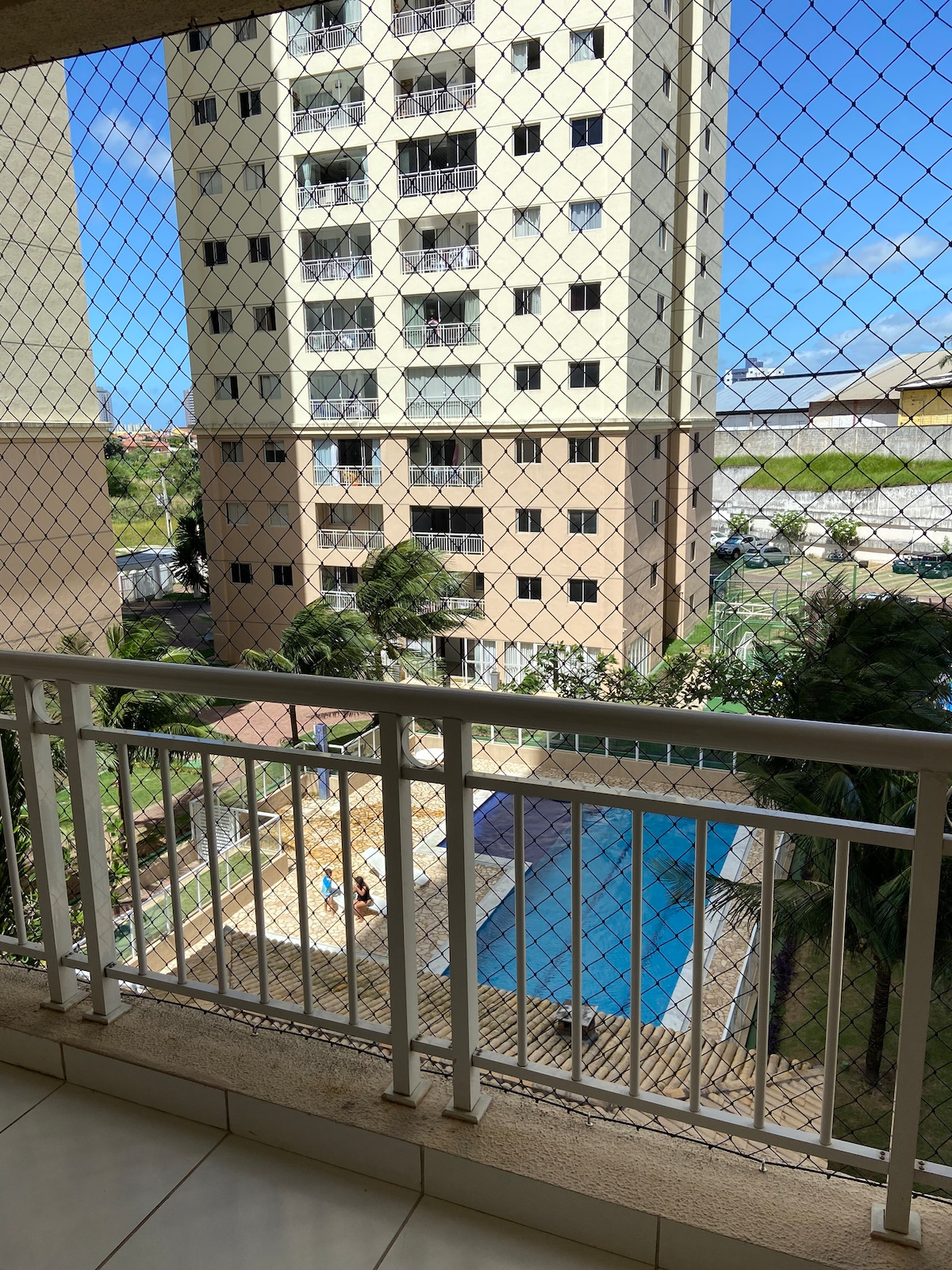 A balcony view offers a glimpse of a shared pool area surrounded by palm trees. The scene features a peaceful atmosphere, with sunlight reflecting off the water. Nearby, a multi-story building stands in the background, framed by clear blue skies.