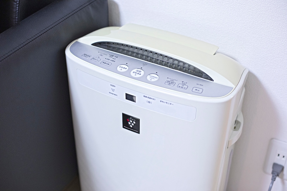 An air purifier is positioned in the corner of the room, featuring a clean, white design. The control panel is visible on the top, displaying various buttons for settings and operation. The device is compact, enhancing the indoor air quality.