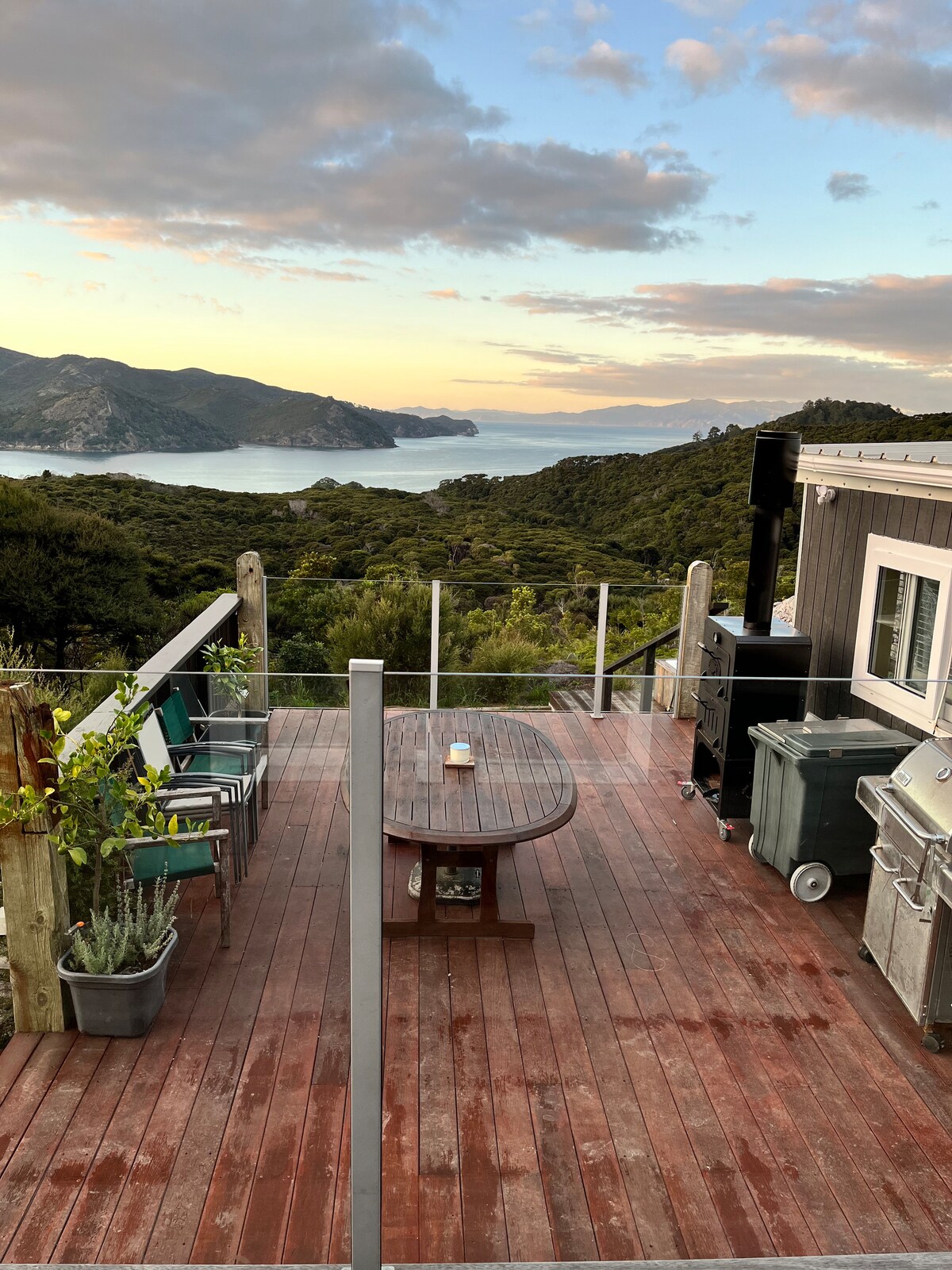 A spacious deck overlooking a scenic coastal view features a round table surrounded by several chairs. Lush greenery and the ocean are visible in the background, complemented by a grill and storage bins nearby. The atmosphere suggests a peaceful outdoor setting.