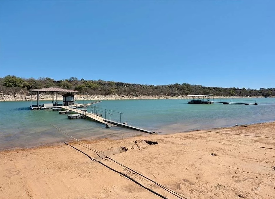 An expansive sandy beach opens up to calm lake waters, featuring two floating docks extending into the lake. Sunlight reflects off the water's surface, while a backdrop of greenery along the shoreline adds a tranquil element.