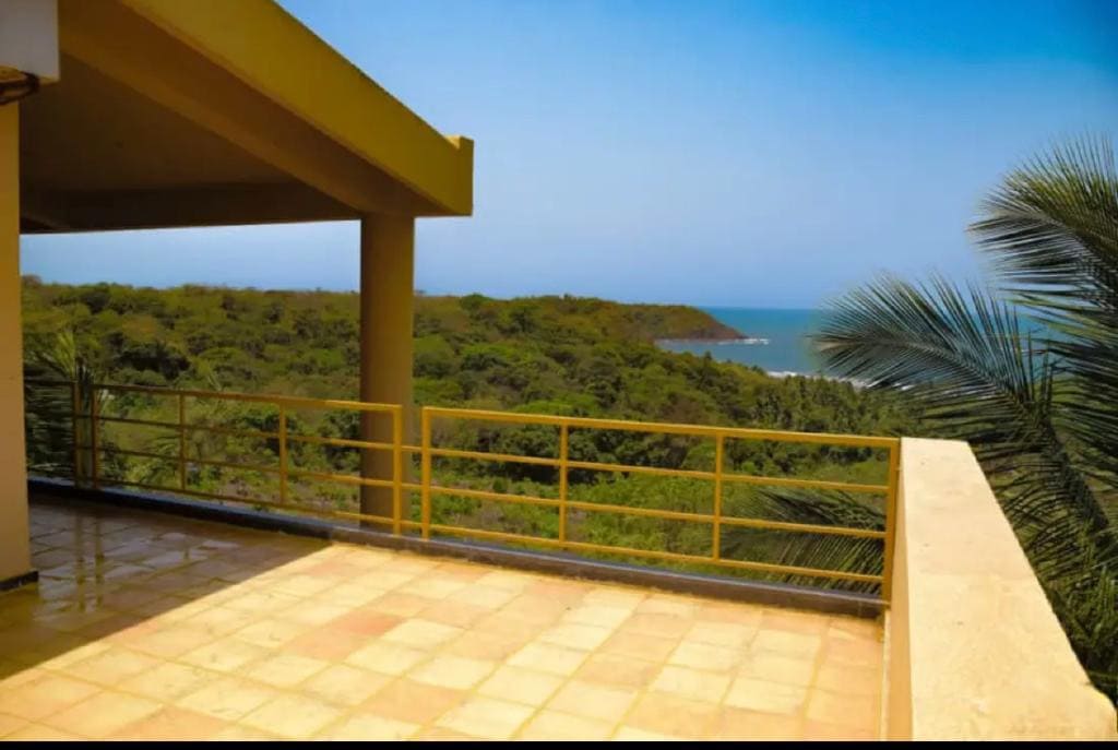A spacious terrace offers expansive views of the ocean and surrounding greenery. A simple railing runs along the edge, complementing the neutral tiled flooring. The clear blue sky and lush landscape create a serene backdrop, making the space ideal for relaxation.