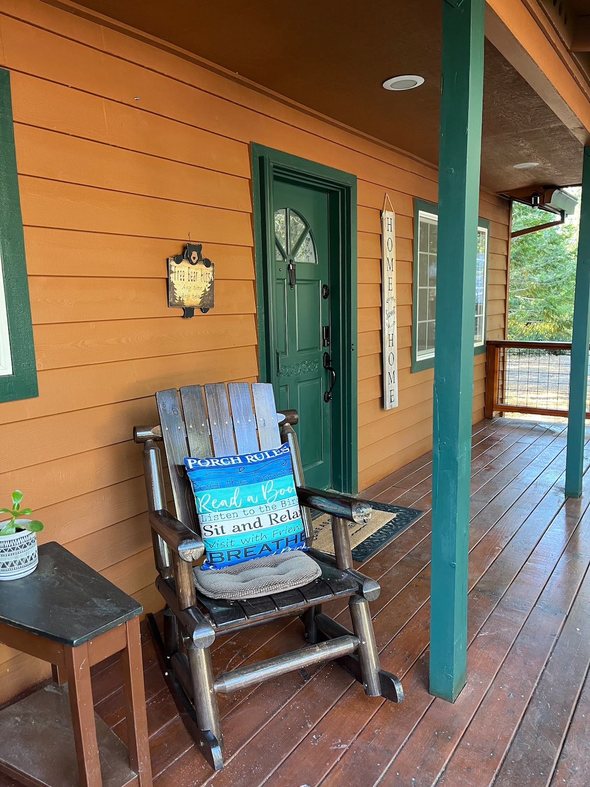 A front porch with wooden flooring features a rustic rocking chair beside a small side table. An inviting green door stands at the entrance, complemented by charming wall decor that enhances the cozy aesthetic of the cabin.