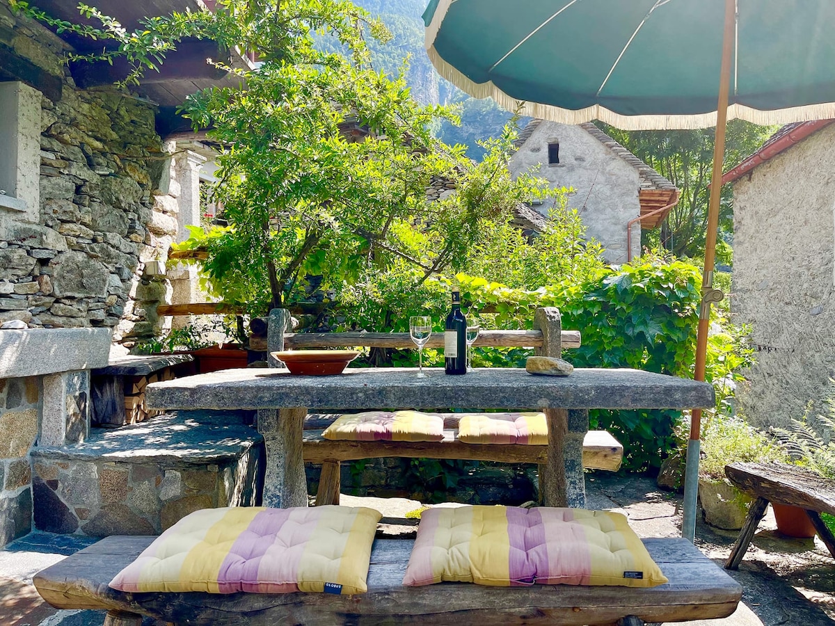 An outdoor seating area features a rustic stone table supported by wooden logs. Two cushioned benches are placed on either side, accompanied by a wine bottle and glasses. Lush greenery surrounds the space, with a large umbrella providing shade.