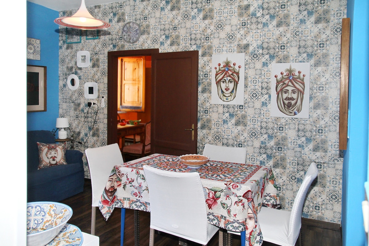A dining area is featured, showcasing a patterned tablecloth over a rectangular table surrounded by white chairs. A blue couch rests against the wall, and decorative plates are displayed on shelves. The walls are adorned with intricate wallpaper and framed art pieces.