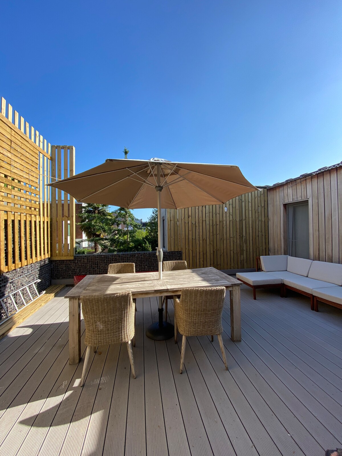 A spacious terrace is shown, furnished with a wooden dining table surrounded by four wicker chairs. A large umbrella provides shade. A cozy seating area is situated to one side, with beige cushions against a wooden backdrop, under a clear blue sky.