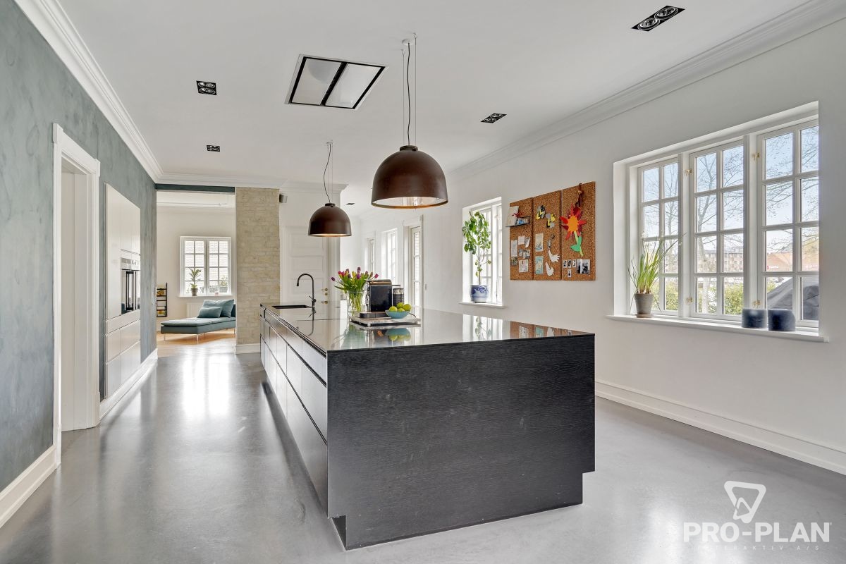 A modern kitchen is showcased, featuring a large central island with a sleek black countertop. Pendant lights hang above, illuminating the space. Windows allow natural light to fill the room, highlighting the simple decor and neutral wall tones that contribute to an open and airy environment.