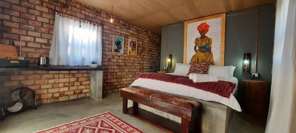 A cozy guest room features exposed brick walls and a spacious bed adorned with white linens and a deep red throw. Artwork of traditional figures enhances the ambiance. A small kitchenette area is visible, accompanied by a wooden bench and decorative rug on the floor.