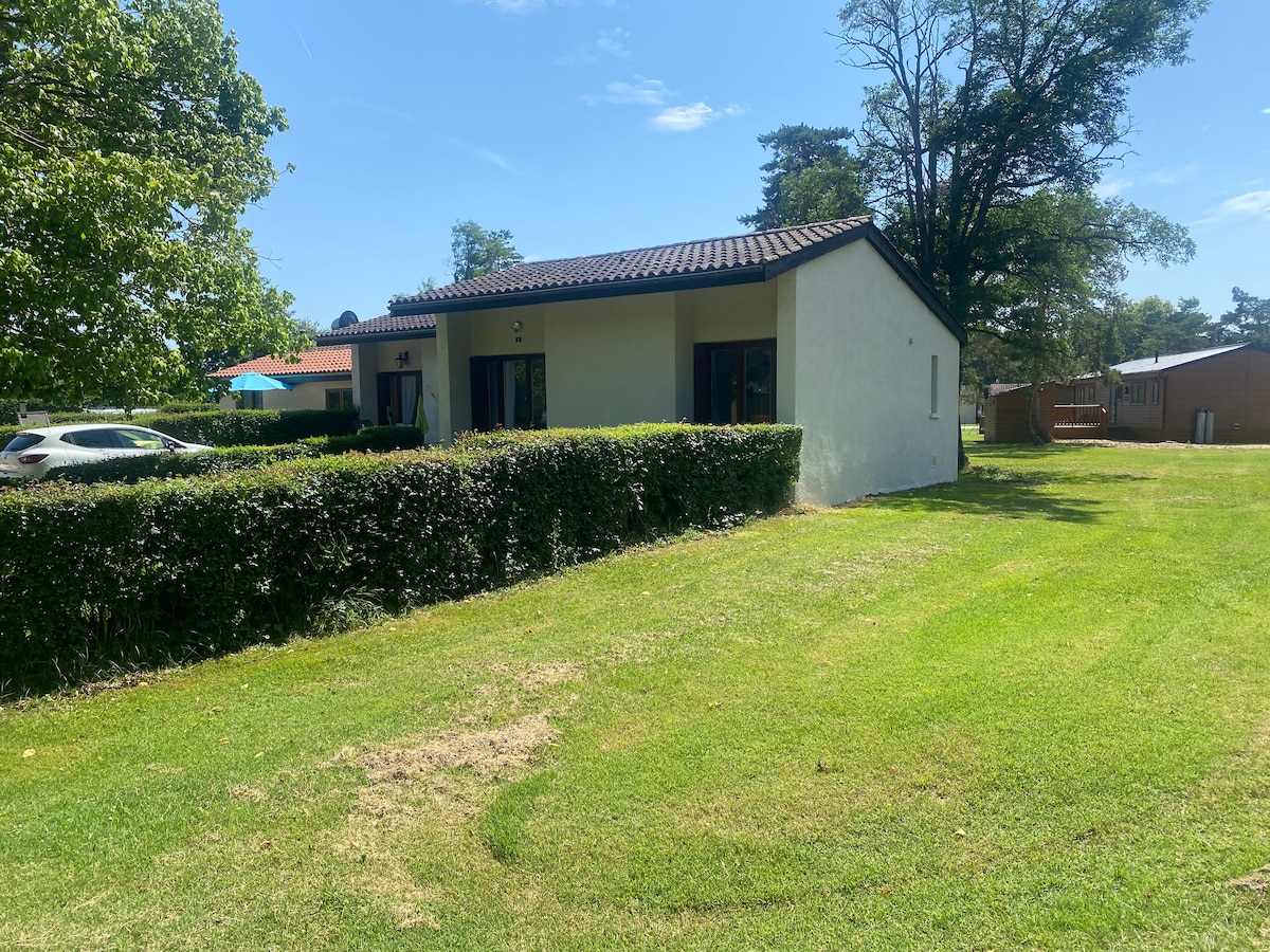 A small bungalow is surrounded by a well-maintained lawn and hedges. The structure features a simple design with a dark tiled roof and multiple windows. In the background, a swimming pool and seating area are partially visible, suggesting outdoor leisure options.