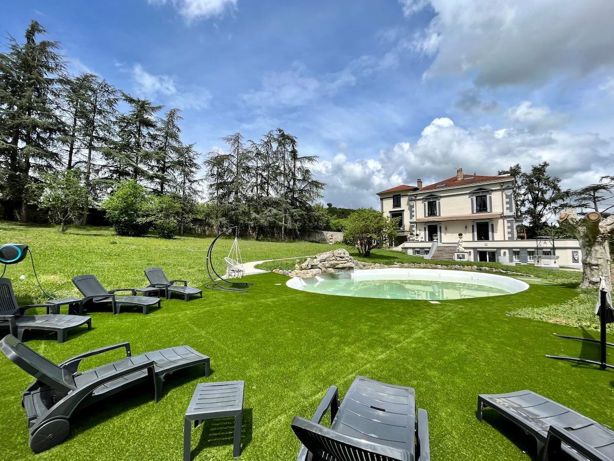 The outdoor area features a manicured lawn with several lounge chairs arranged around a natural-shaped pool. Towering trees provide shade and a hammock offers a relaxed seating option. A large house appears in the background, surrounded by greenery, creating an inviting atmosphere.