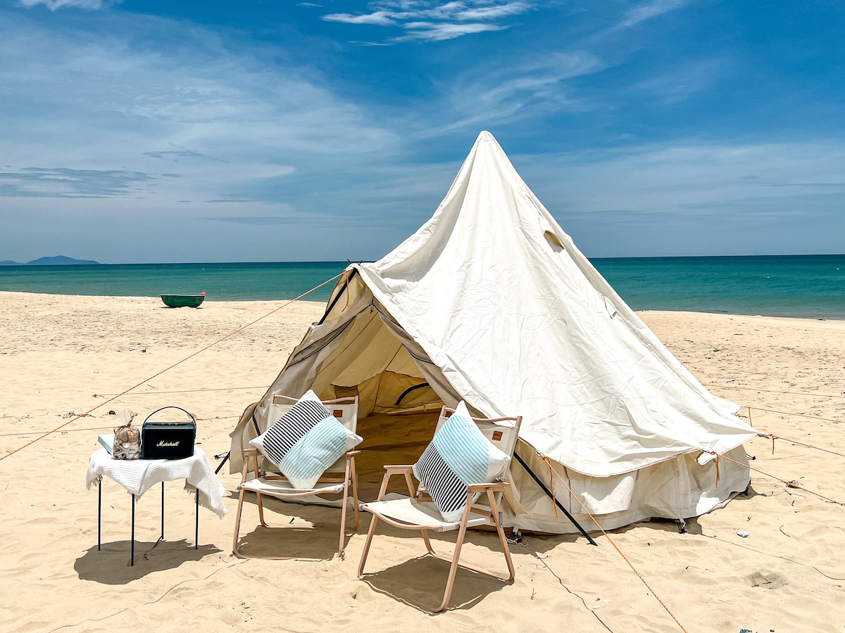A spacious tent is set up on sandy beach, facing a calm sea. Two chairs are positioned nearby, accompanied by a small table that holds a basket. The sky is clear, with a few clouds visible, and a distant boat can be seen on the water.