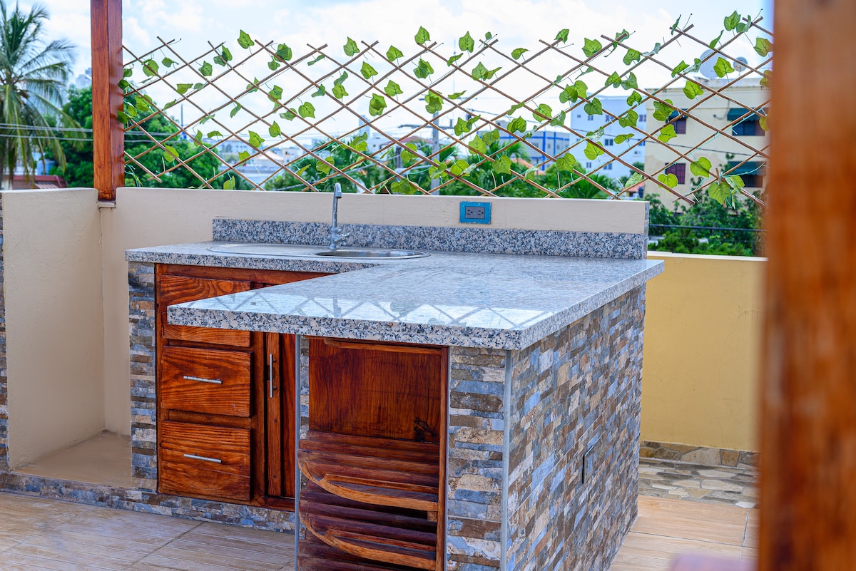 An outdoor kitchen area is showcased, featuring a countertop made of granite and wooden cabinets. A stone wall adds texture, while a lattice above is adorned with climbing plants. The space is bright and open, providing a pleasant atmosphere for outdoor cooking or dining.