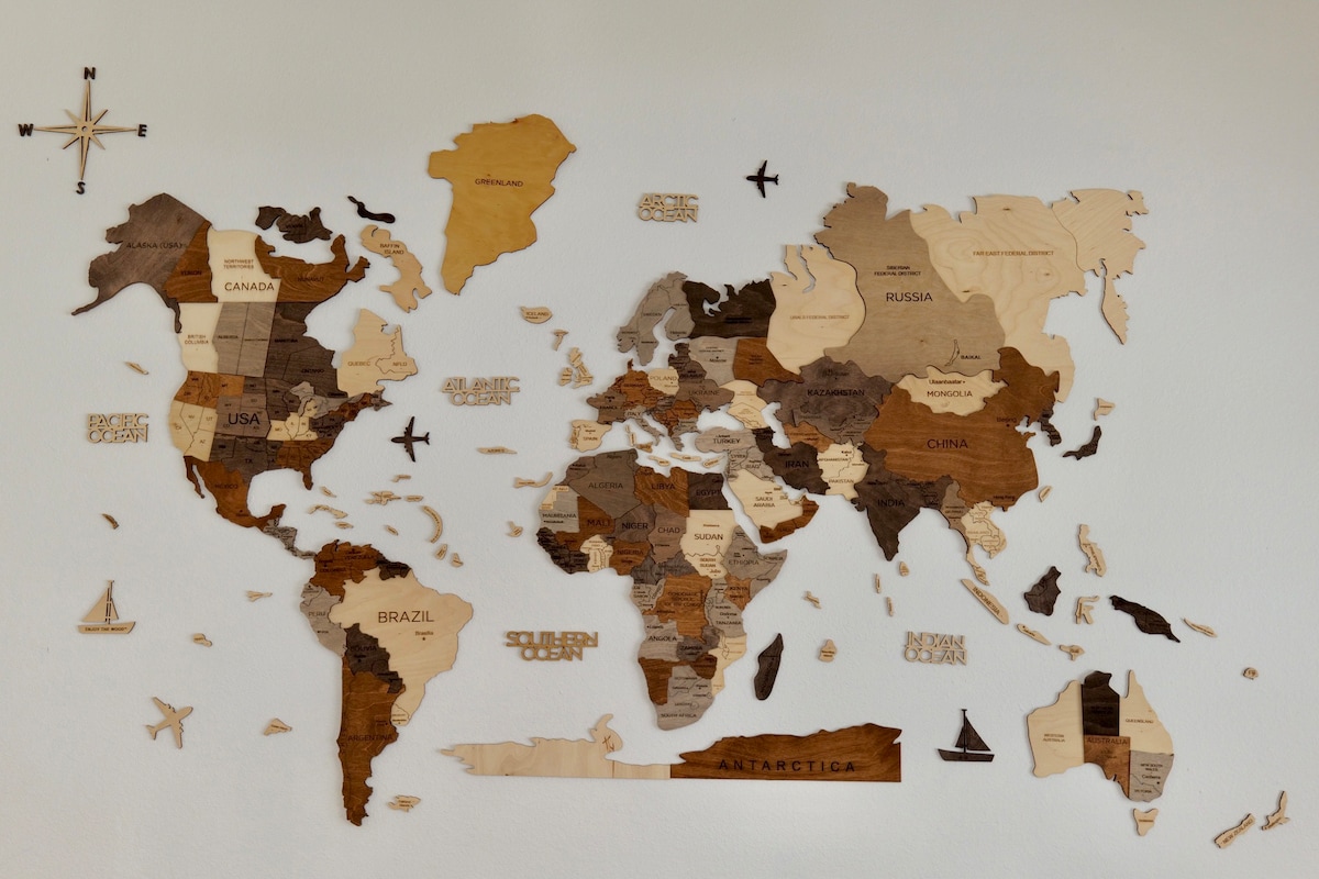 A wooden world map is displayed on a light-colored wall, featuring countries in varying shades of brown and tan. Geographic boundaries are defined, and symbols representing travel, such as airplanes and boats, are incorporated into the design.