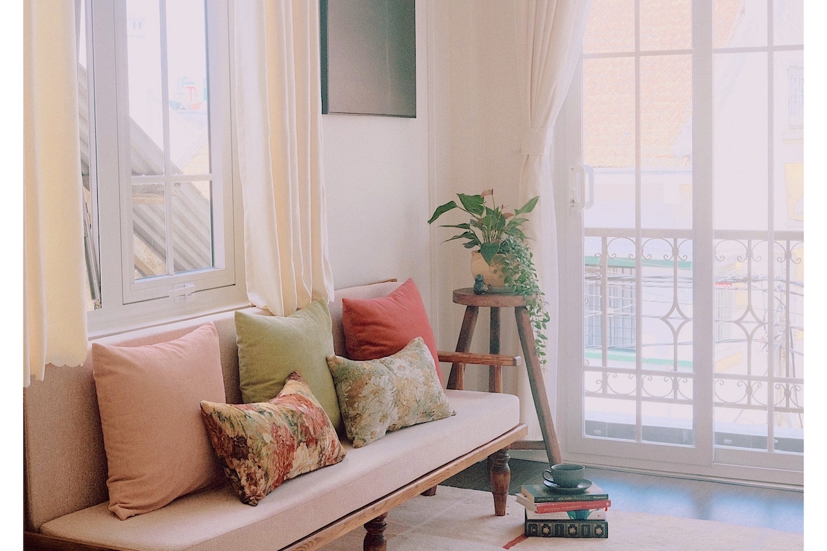 A cozy seating area is presented with a light sofa adorned with decorative pillows in soft hues. A small plant rests on a wooden stand beside the window, which is framed by sheer curtains. Natural light fills the space, creating a warm ambiance.