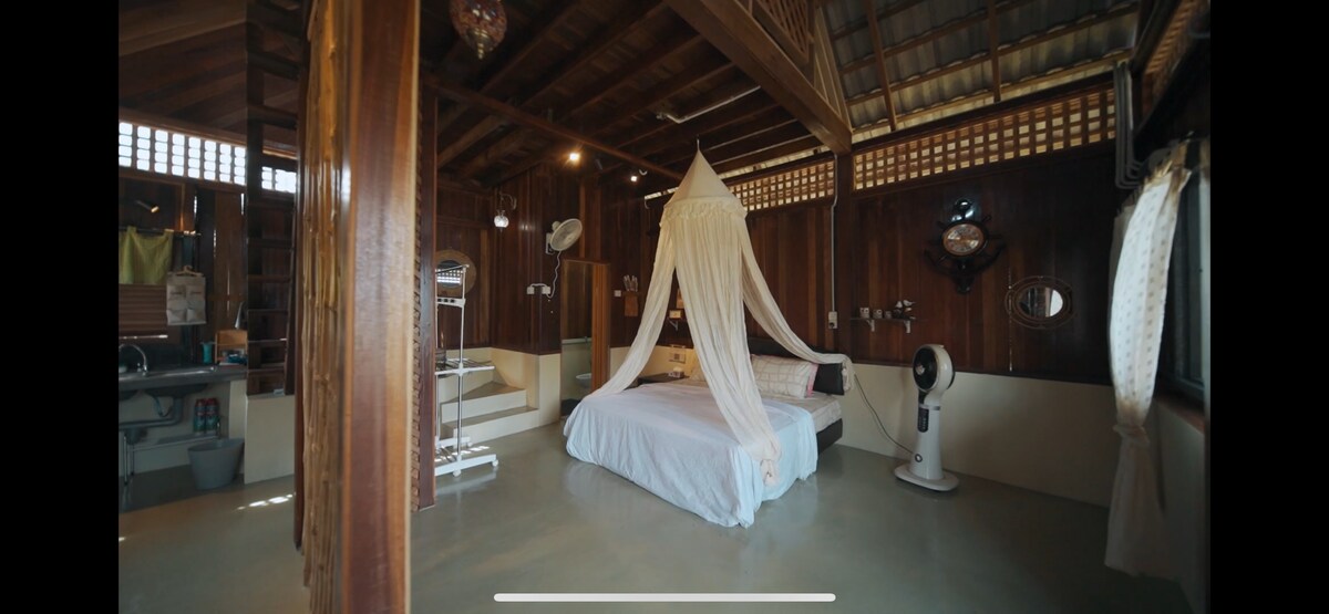 A spacious sleeping area features a large bed draped with a sheer canopy. Wooden walls and ceiling provide a natural ambiance. A fan stands nearby, and soft lighting enhances the tranquil setting. A staircase can be seen in the background, leading to an upper level.