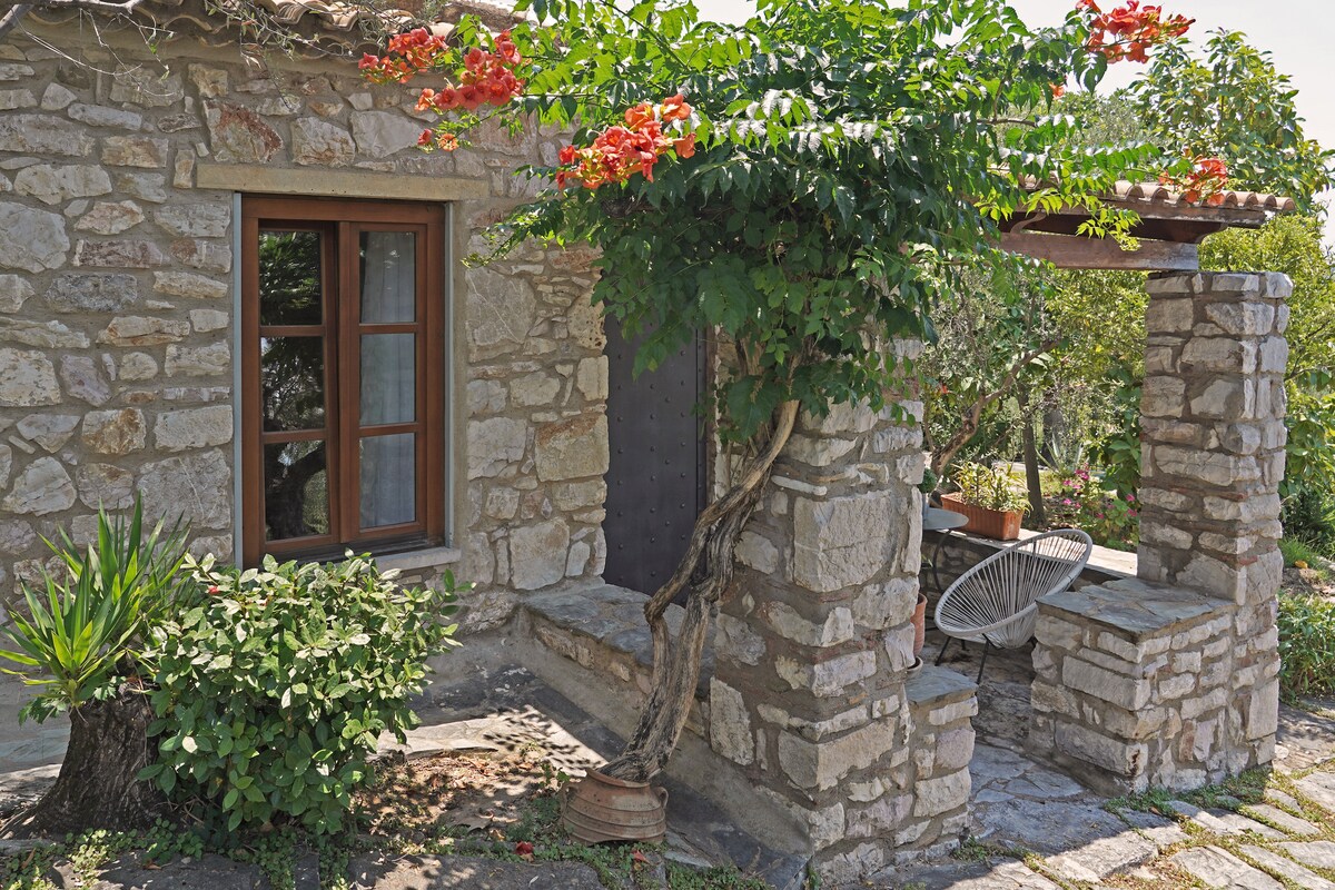 A traditional stone house is featured with wooden window frames and a shaded porch supported by stone columns. Vibrant flowering plants enhance the entrance, while a comfortable chair is positioned nearby. The natural surroundings create a serene setting, inviting relaxation and connection with nature.