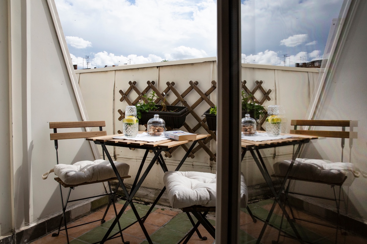 A small outdoor balcony set is visible, featuring a wooden table and two foldable metal chairs. The table is adorned with a glass jar and a drink. Lush greenery grows in a pot nearby, and the sky is partially cloudy, enhancing the atmosphere of the space.