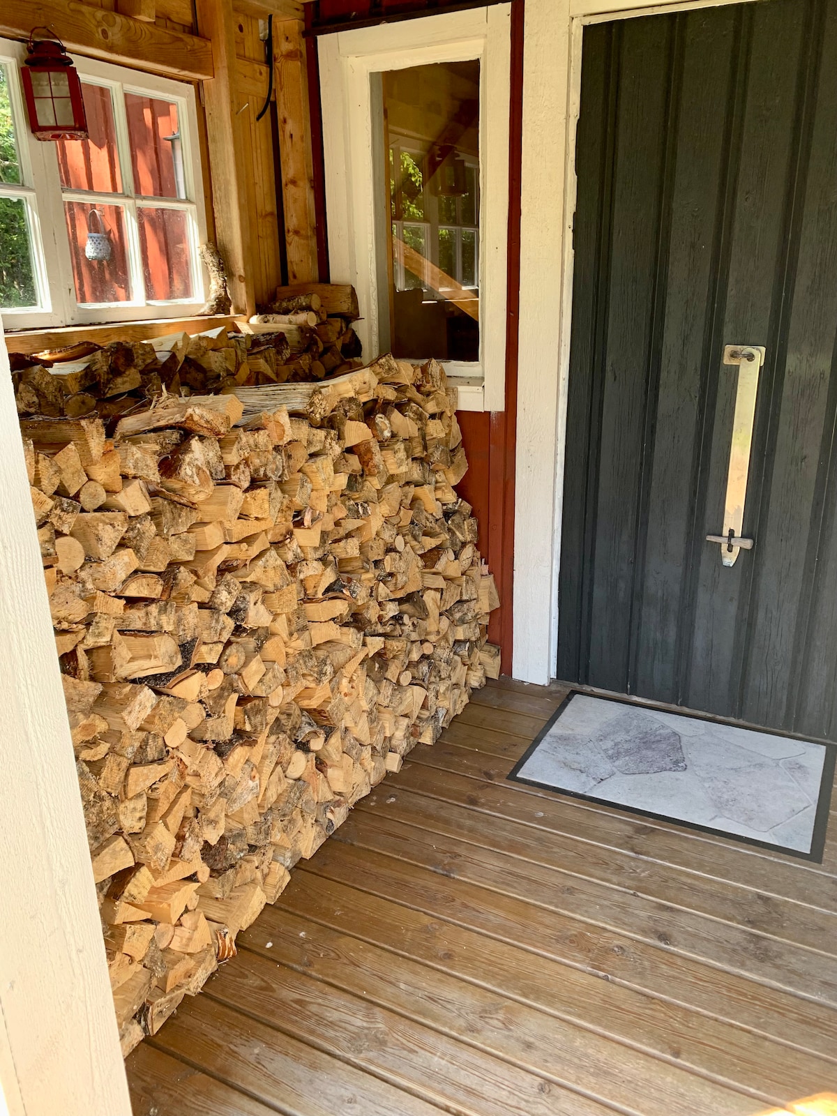 A stack of neatly arranged firewood is positioned against the wall of a wooden porch. The entrance features a dark door and a light-colored mat on the wooden floor, which is surrounded by a rustic wooden structure.