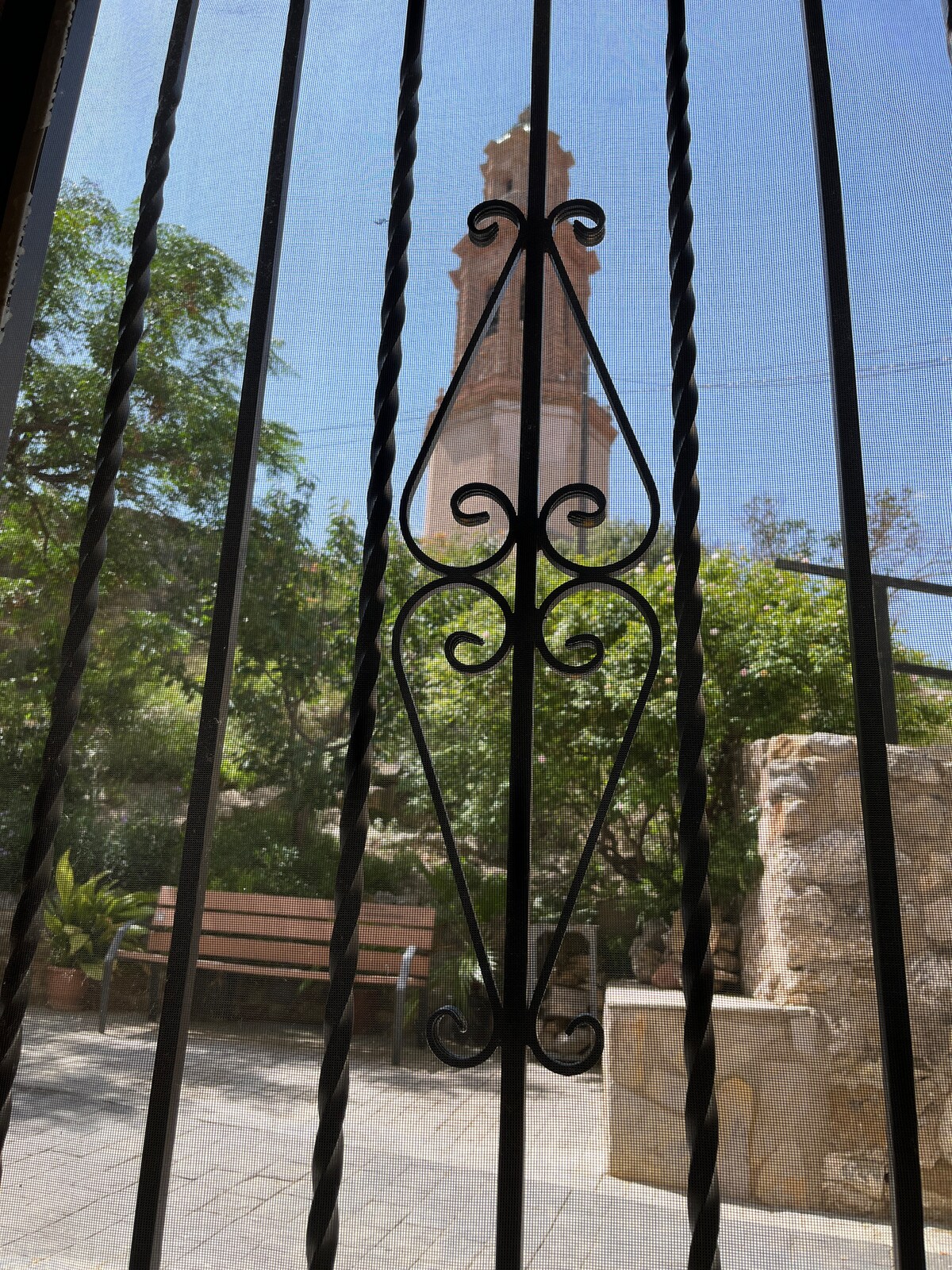 A view is framed by decorative iron bars, showcasing a nearby Mudejar tower. Lush greenery and a stone bench are visible in the foreground, enhancing the quaint atmosphere. The sunlight filters through, highlighting the textures of the stone and plants.