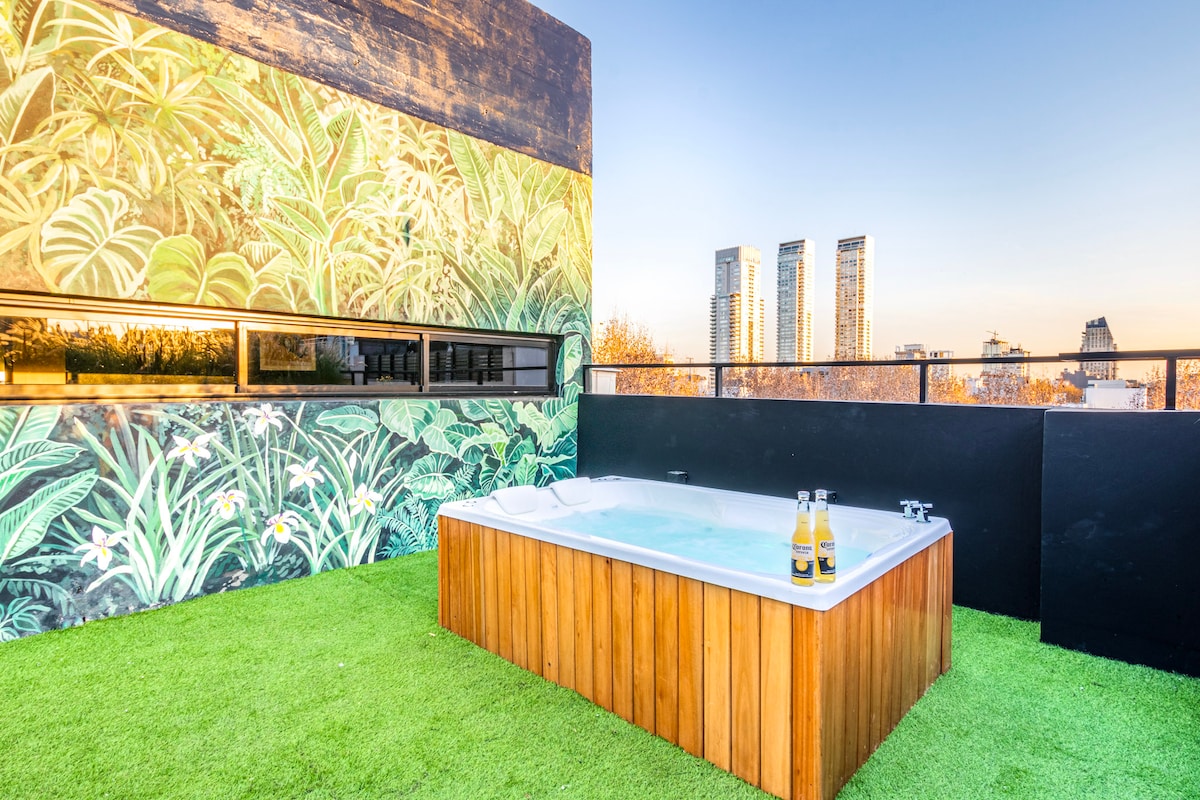 A rooftop patio features a wooden jacuzzi surrounded by lush green grass. An artistic mural of tropical plants adds a vibrant backdrop. City skyscrapers can be seen in the distance, under a clear sky that transitions into the warm hues of sunset.