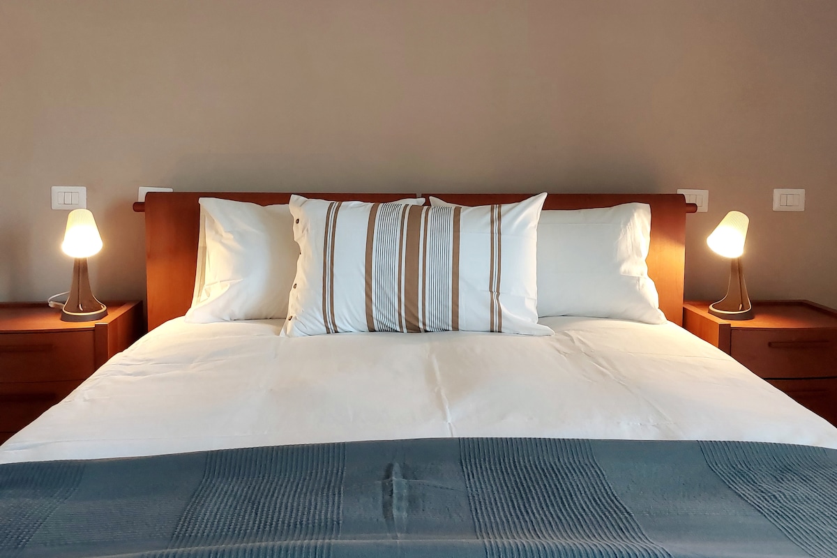 A spacious bed with crisp white linens is flanked by two bedside tables, each featuring a softly glowing lamp. Decorative pillows add a touch of elegance to the bed, set against a warm, neutral wall.
