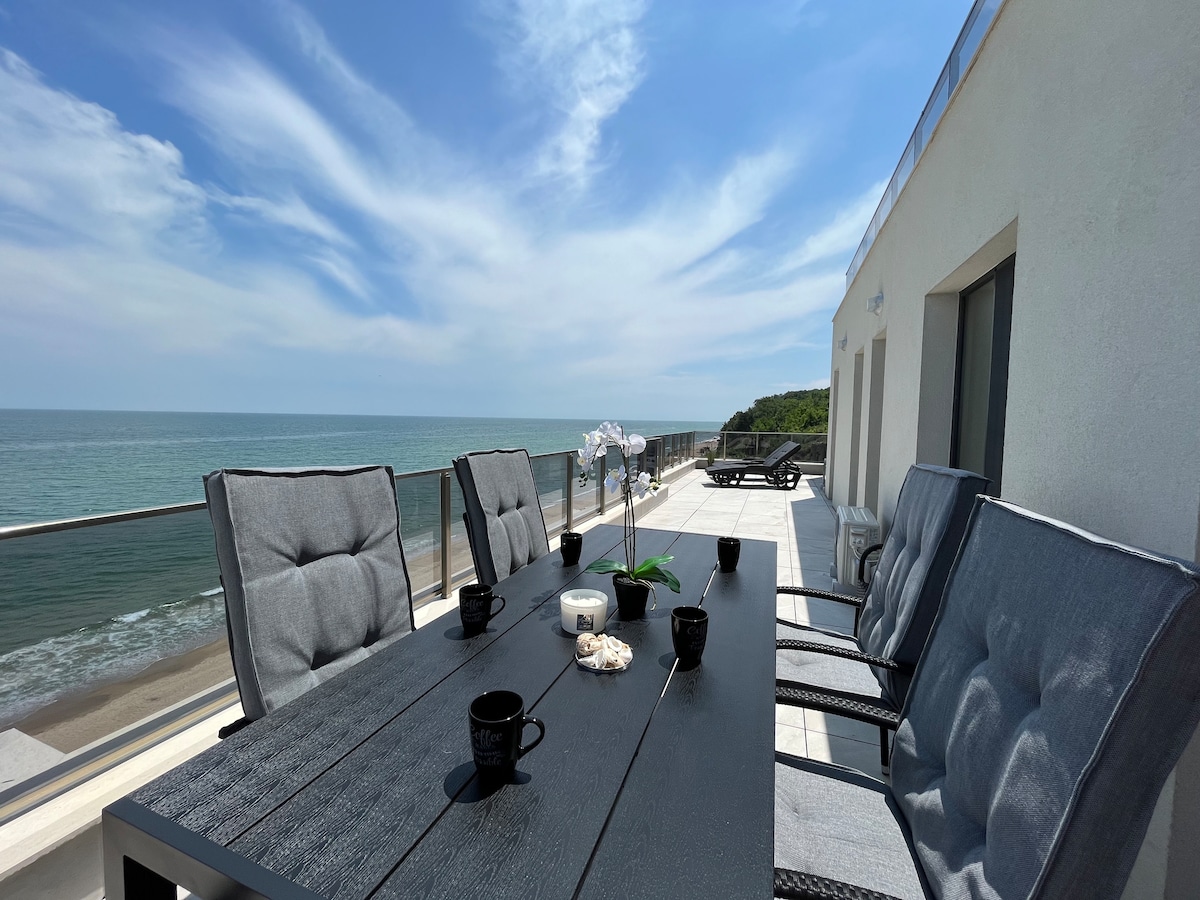 A large terrace is shown with a black dining table surrounded by comfortable gray chairs. Cups and a decorative arrangement are present on the table. The tranquil sea and sky create a serene backdrop, emphasizing the space's coastal atmosphere.