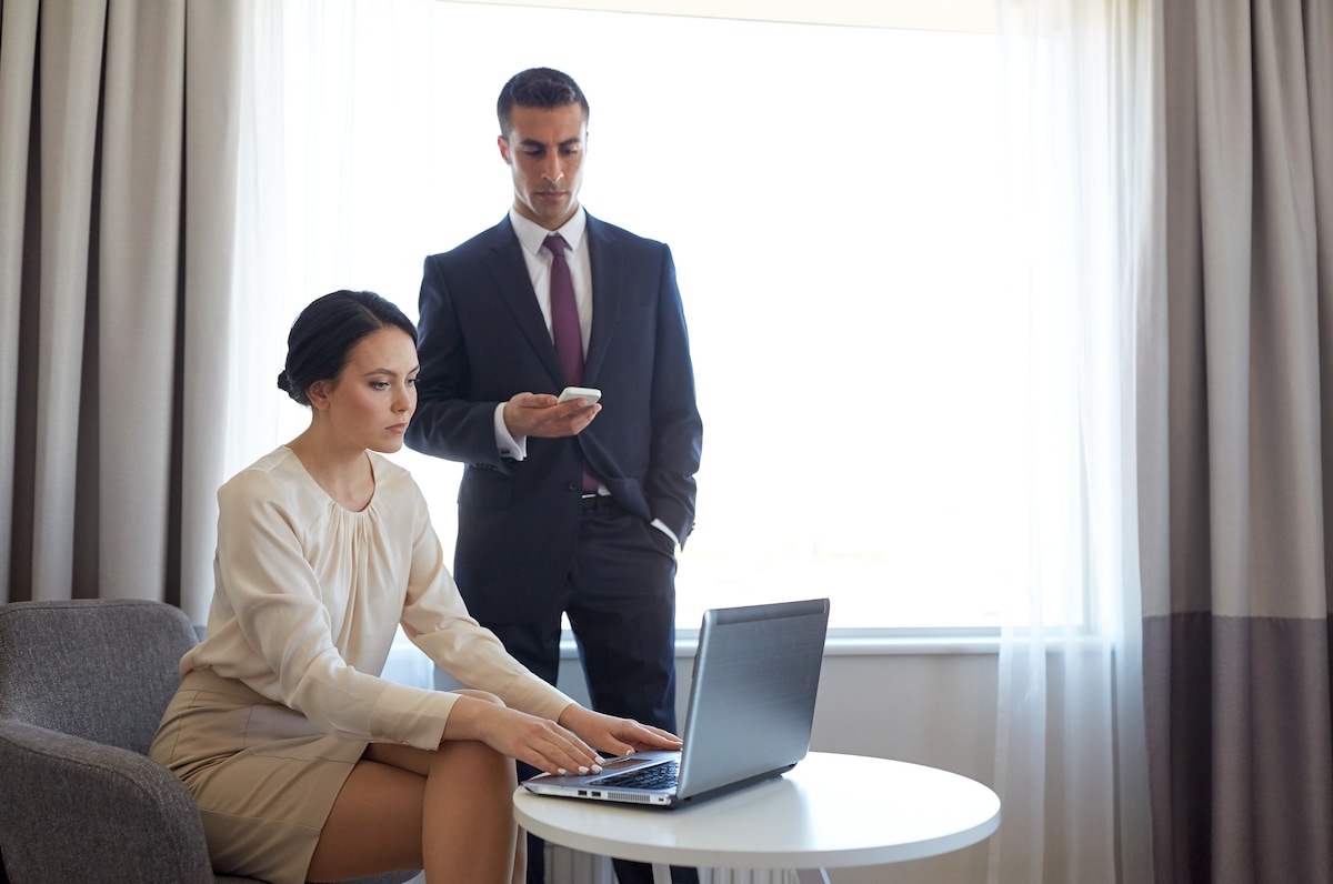 A modern workspace is presented with a sleek laptop on a round table. A professional suit is worn by a standing figure, while a seated individual types. Soft curtains adorn a large window, allowing natural light to brighten the space.