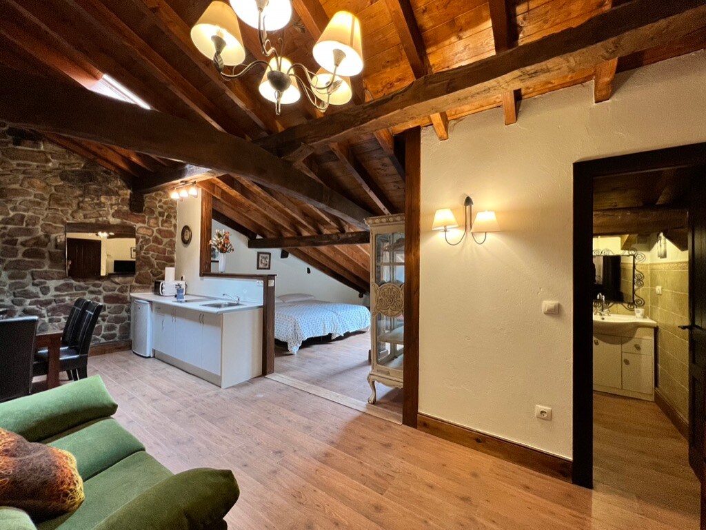 An open living space features wooden beams overhead, creating a rustic feel. A dining area includes a table and chairs, while a kitchenette is visible. A spacious bed is located in a separate nook, with a bathroom entrance seen in the background. Natural light enters through the windows.