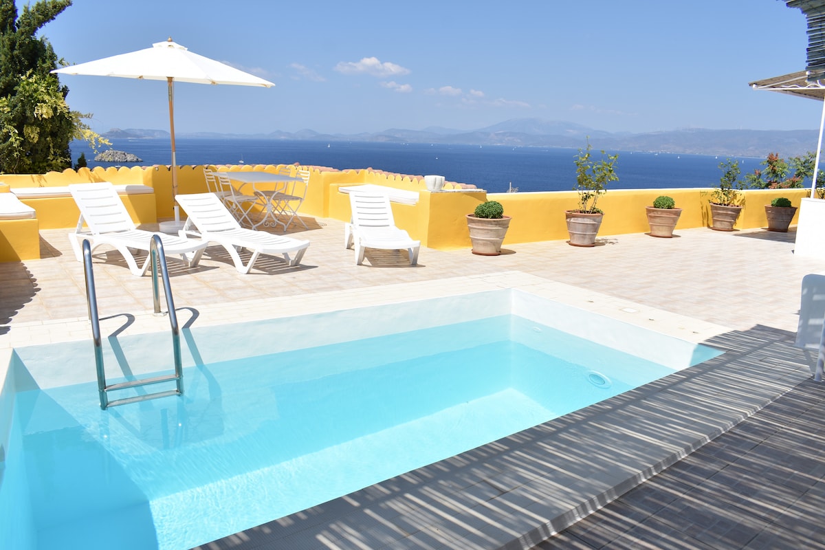 A private swimming pool is surrounded by sun loungers under an umbrella on a spacious terrace. Potted plants add greenery to the yellow-walled area, with stunning views of the blue gulf and distant mountains visible in the background under a clear sky.