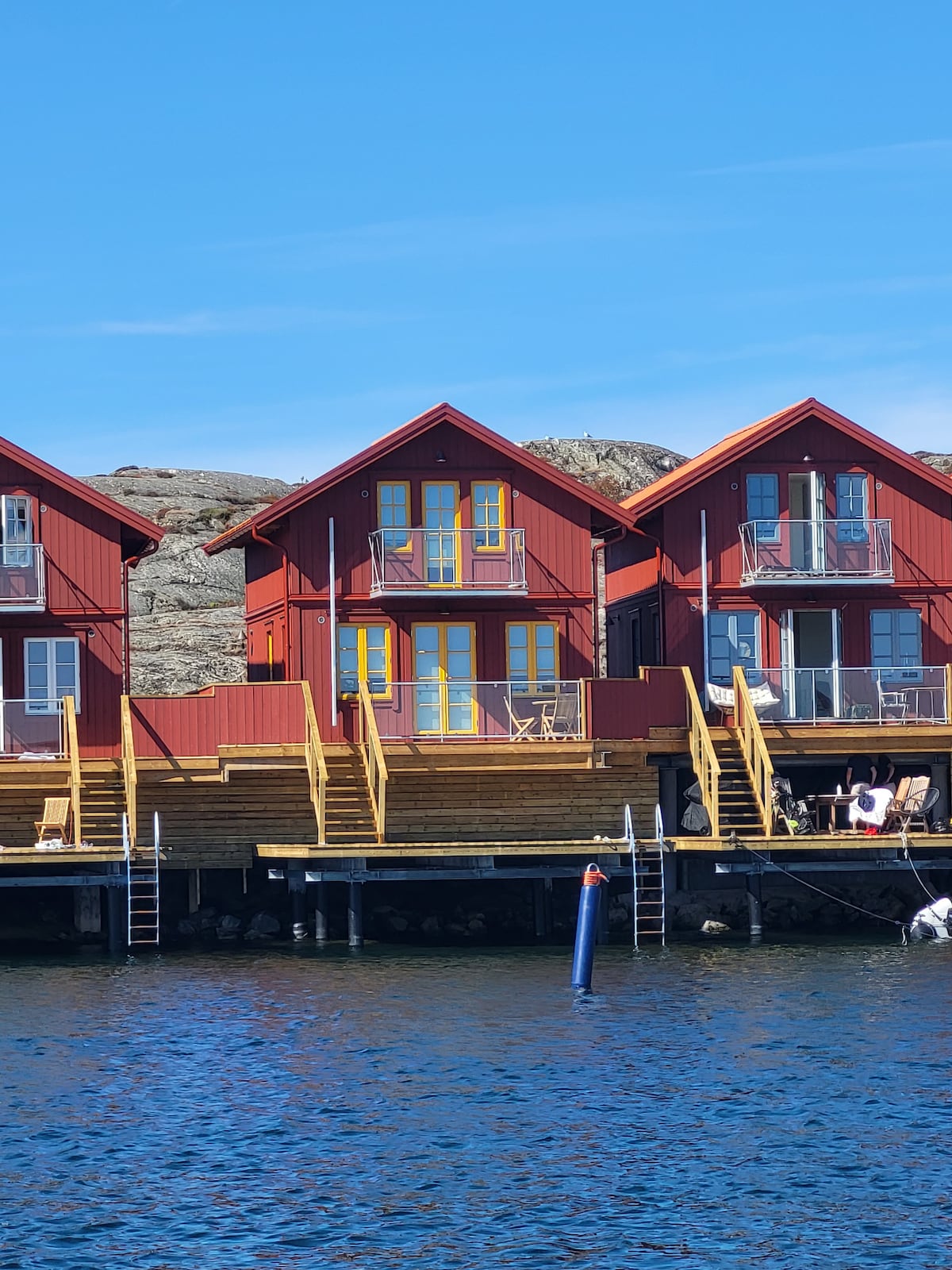 A charming row of newly built cottages is positioned along the water's edge. Each cottage features a balcony with seating, large windows reflecting sunlight, and is accessed by wooden staircases. The surrounding rocky landscape is visible against a clear blue sky.