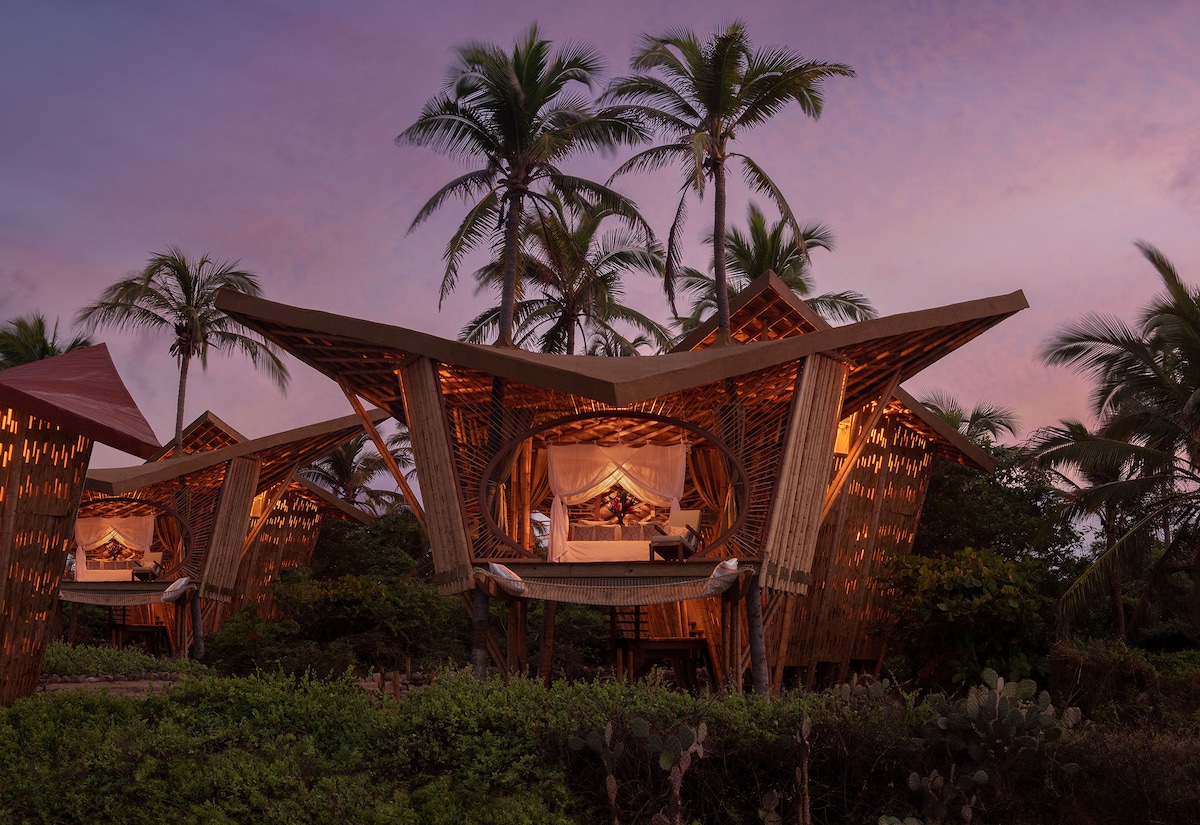 A unique treehouse structure is surrounded by palm trees, showcasing its architectural design with bamboo elements. Soft lighting adds warmth to the cozy sleeping area, where a spacious bed is visible, set against a backdrop of the evening sky.