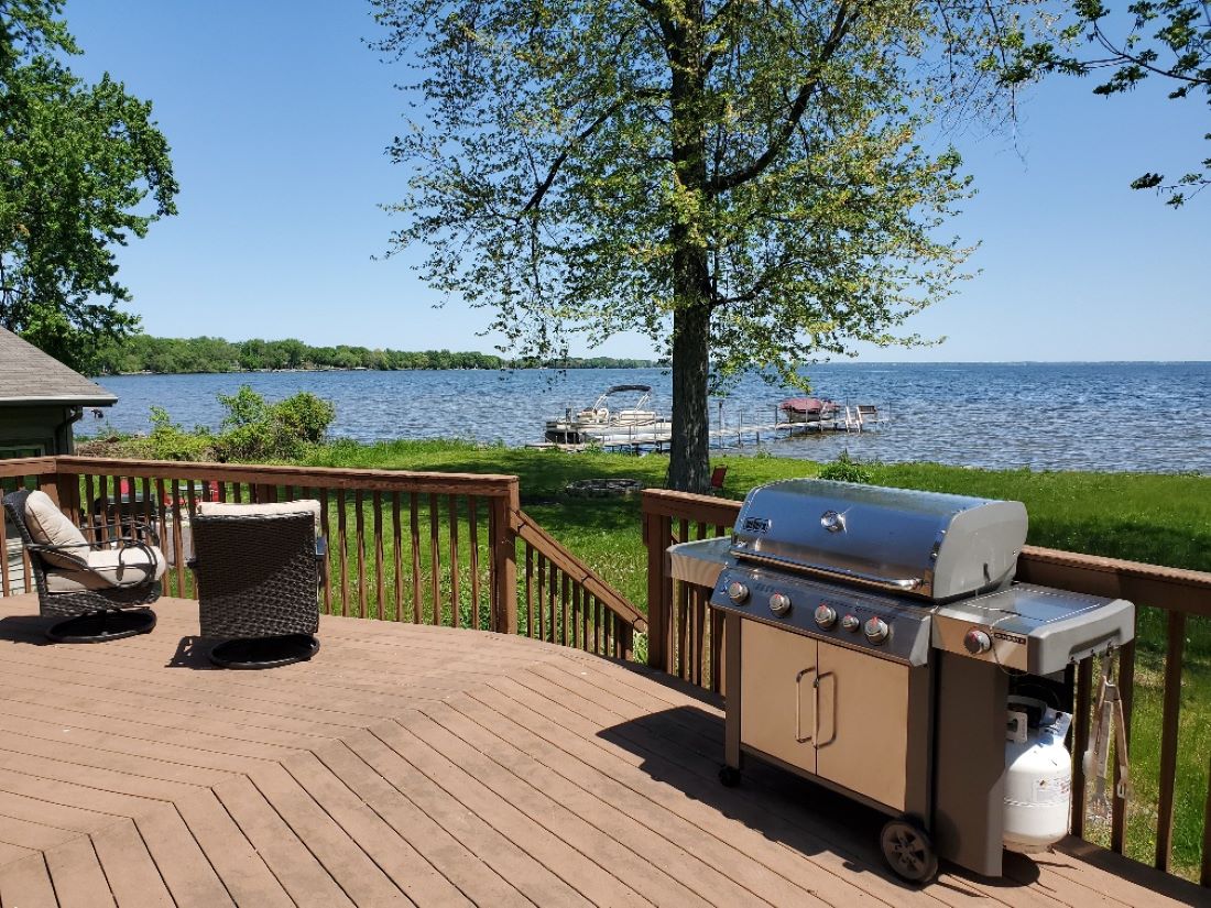 A spacious cedar deck features a modern grill, inviting outdoor cooking. Two comfortable chairs provide seating and a view of the lake. Vibrant green grass surrounds the area, and the serene water stretches into the distance under a clear blue sky.