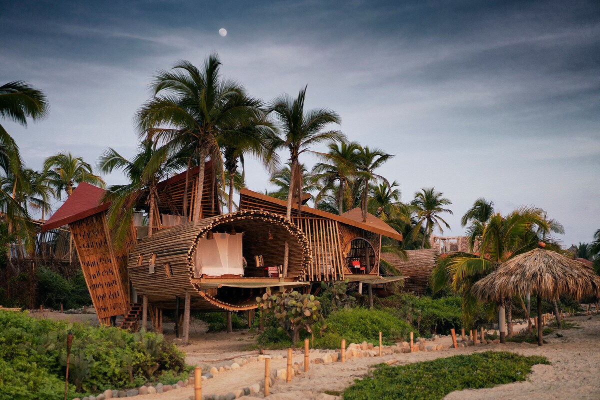 A uniquely designed treehouse with a conical shape is surrounded by palm trees, positioned near sandy terrain. The structure features large openings that provide views of the outdoor environment, while a cozy sitting area is partially visible on the porch.