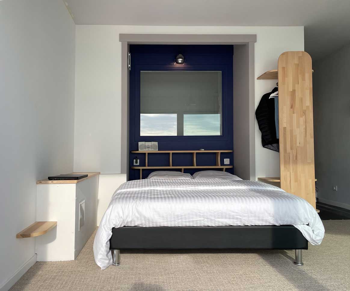A serene sleeping area features a spacious bed with white linens. A large window framed in navy blue allows ample natural light, while a light wood shelving unit provides storage space. Additional shelving is placed above the bed, enhancing the functionality of the room.