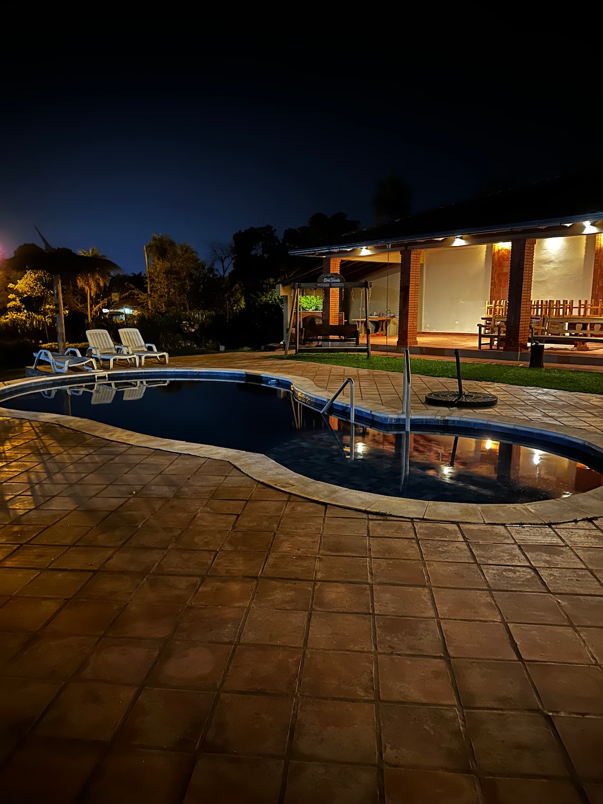 An inviting outdoor area is highlighted by a softly lit swimming pool surrounded by tiled flooring. Lounge chairs are positioned nearby, while a covered seating area is visible in the background. Subtle lighting enhances the serene ambiance of the evening setting.