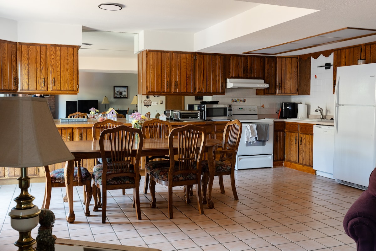 A spacious kitchen and dining area is featured with wooden cabinetry and tiled floors. A large dining table with several chairs sits prominently, while various kitchen appliances are visible along the counters. Warm lighting enhances the inviting feel of the room.