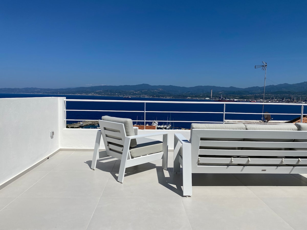 A rooftop terrace features modern white lounge chairs arranged around a small coffee table. Expansive sea views are visible alongside distant mountains, while a clear blue sky enhances the serene atmosphere.