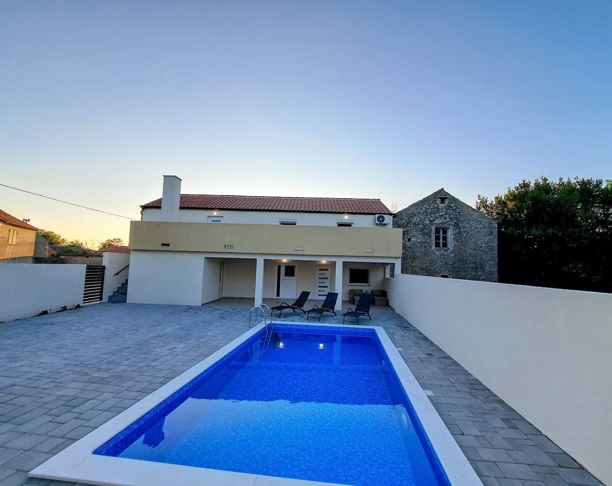 The exterior of the house is captured under a clear sky, featuring a swimming pool surrounded by a spacious patio. Several lounge chairs are placed beside the pool, and the two-story house is designed with a minimalist facade, complemented by a traditional stone building in the background.