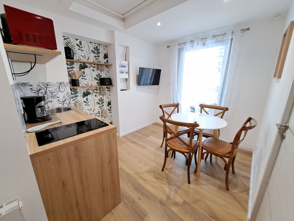 A modern kitchen area features wooden cabinetry with an electric stovetop and coffee maker. A round dining table with four rattan chairs is positioned nearby. A wall shelf with decorative items and a wall-mounted television can be seen. Natural light filters in through a window with light curtains.