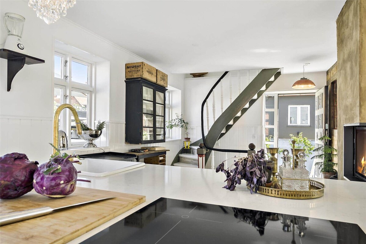 A spacious kitchen area is highlighted by sleek countertops and modern appliances. A wooden cutting board with fresh produce is placed in the foreground. The surrounding decor includes a charming spiral staircase and large windows, allowing natural light to fill the space.