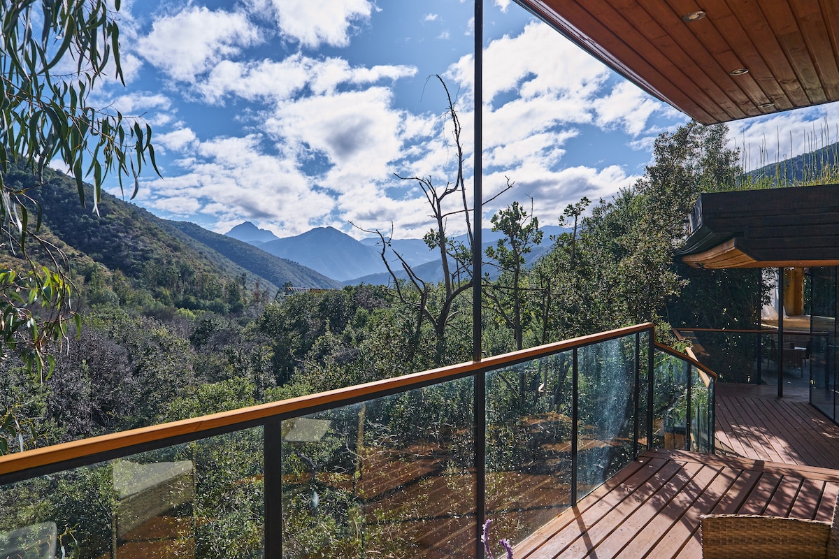 A spacious wooden deck features glass railings, offering unobstructed views of a lush valley surrounded by mountains. The sky is filled with soft clouds, complementing the natural green landscape below.