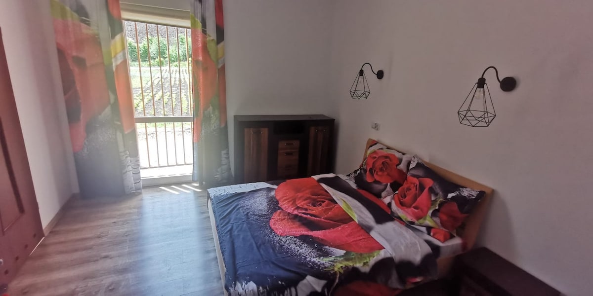 A cozy room features a spacious bed adorned with vibrant floral bedding. Natural light fills the space through a set of glass doors, leading to an outdoor area. Two decorative light fixtures hang above, and a wooden dresser is positioned nearby.