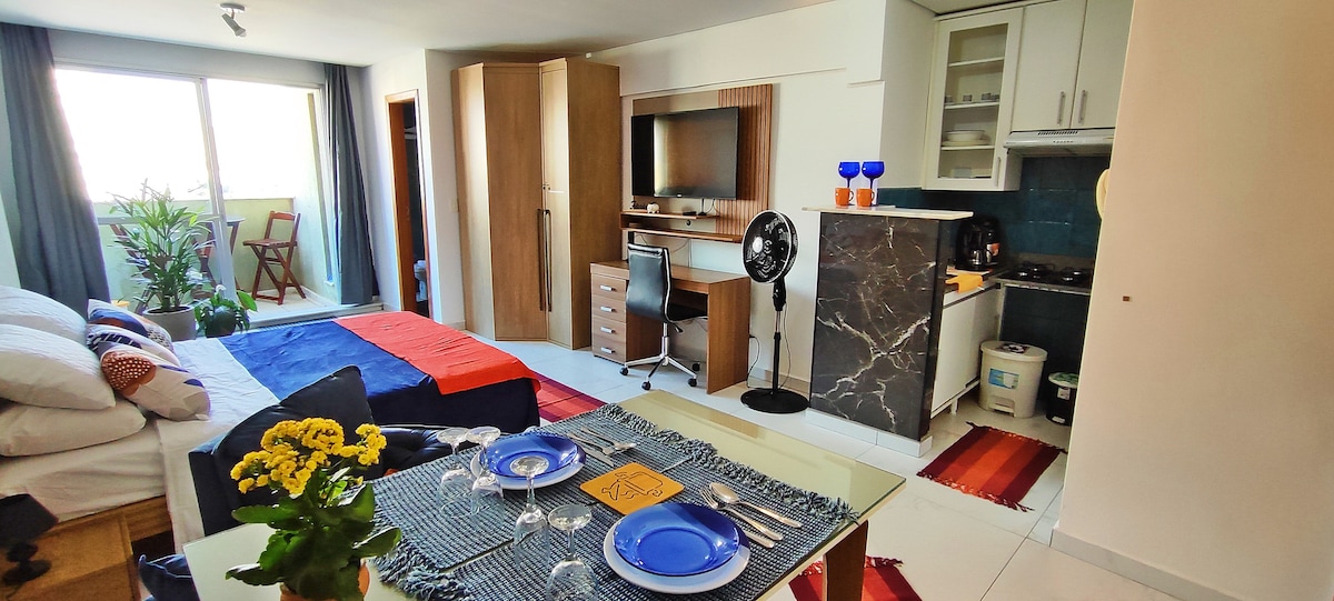 A modern loft interior showcases a combination of living and dining spaces. A neatly arranged table with blue plates sits in front of a colorful woven placemat. The kitchen area includes cabinets, a stove, and a small refrigerator, while large windows allow natural light to enter.